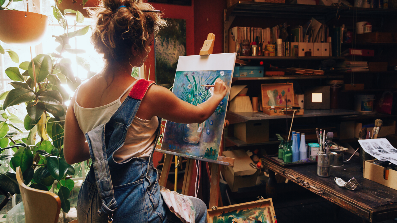 A person sits in a studio and paints a vase and flowers on a canvas.