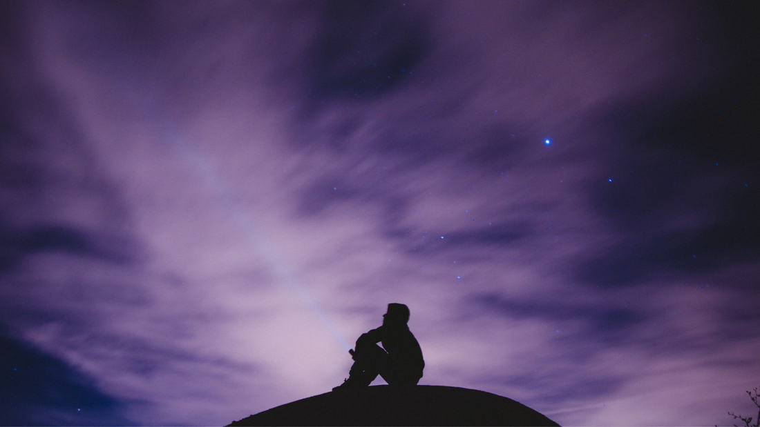A shadow of a person sits on a hill below a purple sky, reflecting on life.